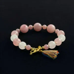 A top-down view of a beaded crystal bracelet featuring varying sizes of translucent Pink Quartz, purple Ametrine, and clear White Ghost Crystal beads. The bracelet is finished with a polished gold toggle clasp and a vibrant multi-color thread tassel.