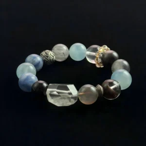 A top-down photograph of a mixed gemstone beaded bracelet on a black background. The bracelet features prominent pale blue Aquamarine rounds, a clear faceted Green Phantom Quartz bead with green inclusions, along with clear quartz, black protective stones, chatoyant labradorite, and patterned silver spacer beads. It is designed for Feng Shui balance and chakra healing.