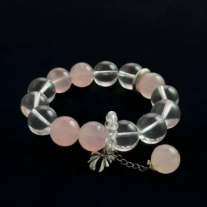 A close-up photograph of a gemstone bracelet featuring alternating round beads of transparent Clear Quartz and translucent pink Rose Quartz. It is finished with a small silver metal bow charm featuring a dangling pink quartz bead, set against a black background.