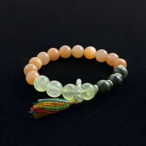 A top-down view of a beaded Feng Shui crystal bracelet on a black background. The bracelet features peach-colored Orange Moonstone beads, translucent Green Rutilated Quartz beads with dark inclusions, dark green chatoyant Seraphinite (Clinochlore) beads, and a multi-colored thread tassel attached near clear quartz guru beads. It is designed for heart and sacral chakra healing.